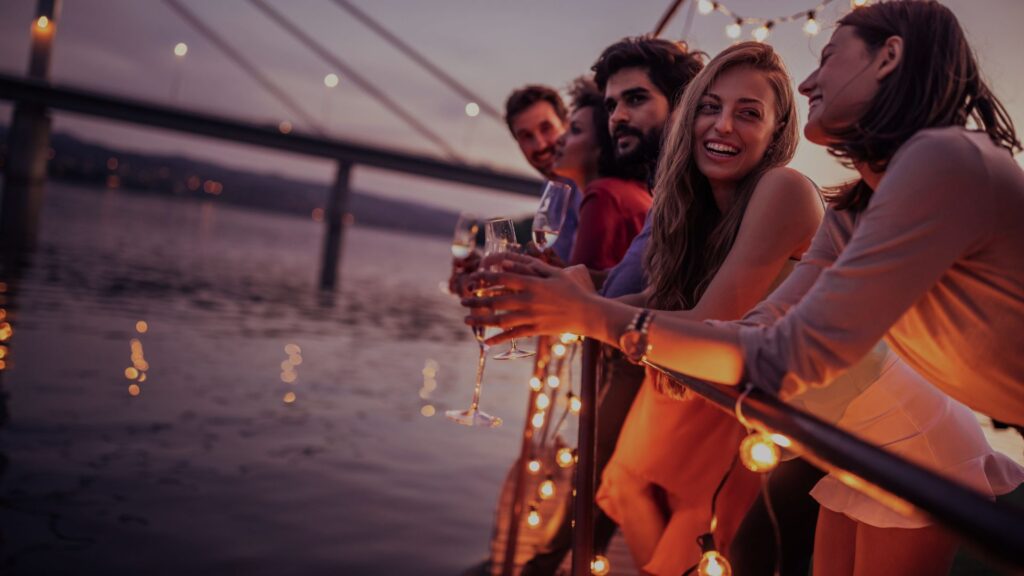 Gruppe von vier Freunden, die bei Sonnenuntergang am Wasser sitzen und Wein trinken.