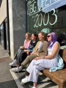 Vier Frauen sitzen auf einer Bank vor einem Café, lachen und trinken Kaffee.