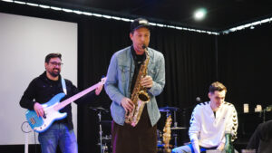 Drei Musiker auf einer Bühne, einer spielt Saxophon, die anderen Gitarre und Bass.