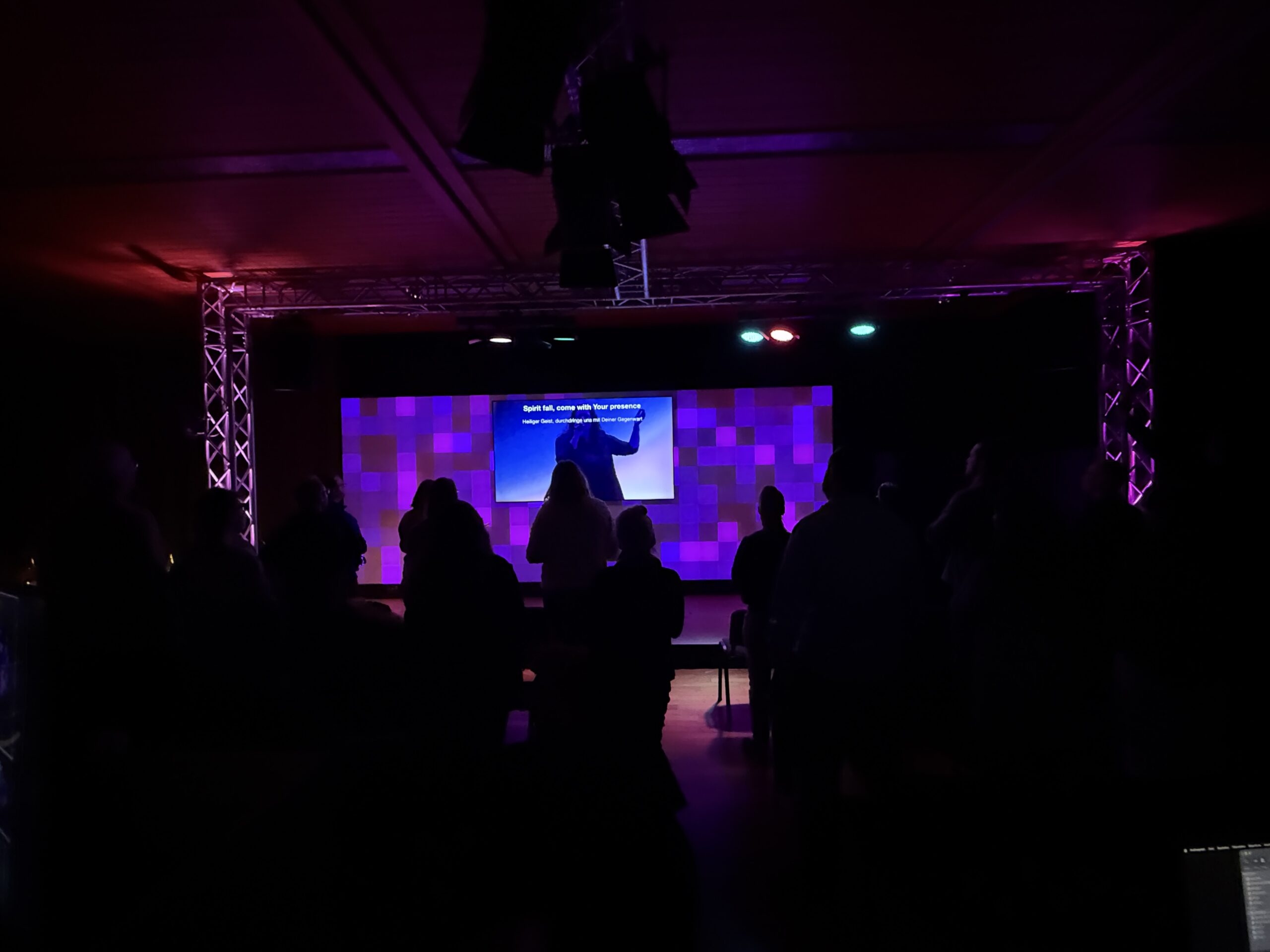 Dunkler Raum mit mehreren silhouettenhaften Personen vor einem großen Bildschirm, der blaue und pink.