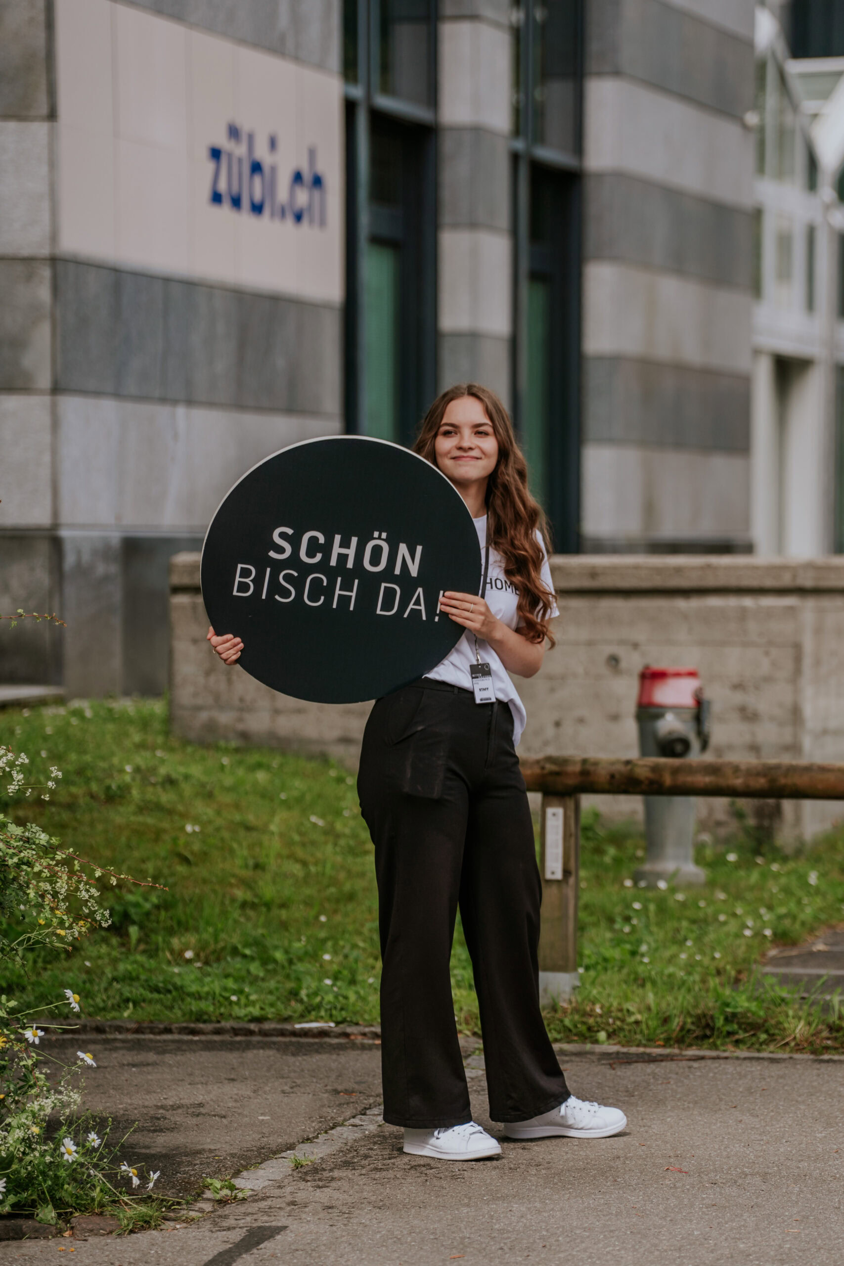 Frau steht draußen vor einem Gebäude, hält ein rundes Schild mit der Aufschrift "Schön, dass du da b.