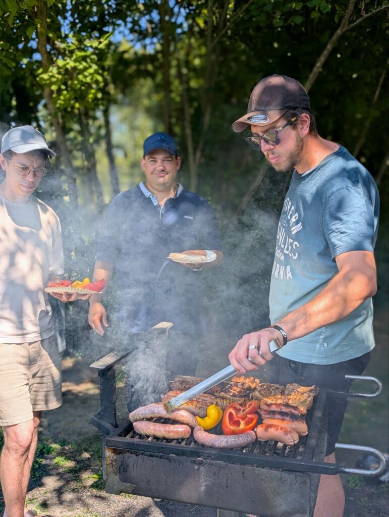 Trois hommes autour d’un barbecue en plein air, cuisson de viande et légumes.