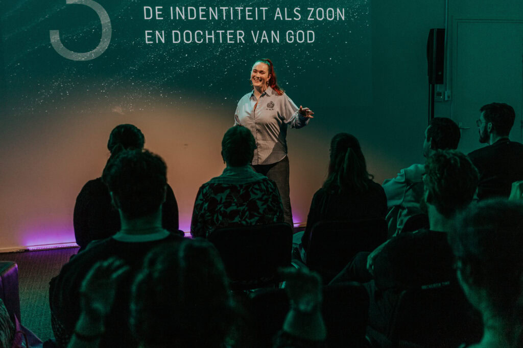 Vrouw staat op podium en spreekt voor een groep mensen in een donkere ruimte.