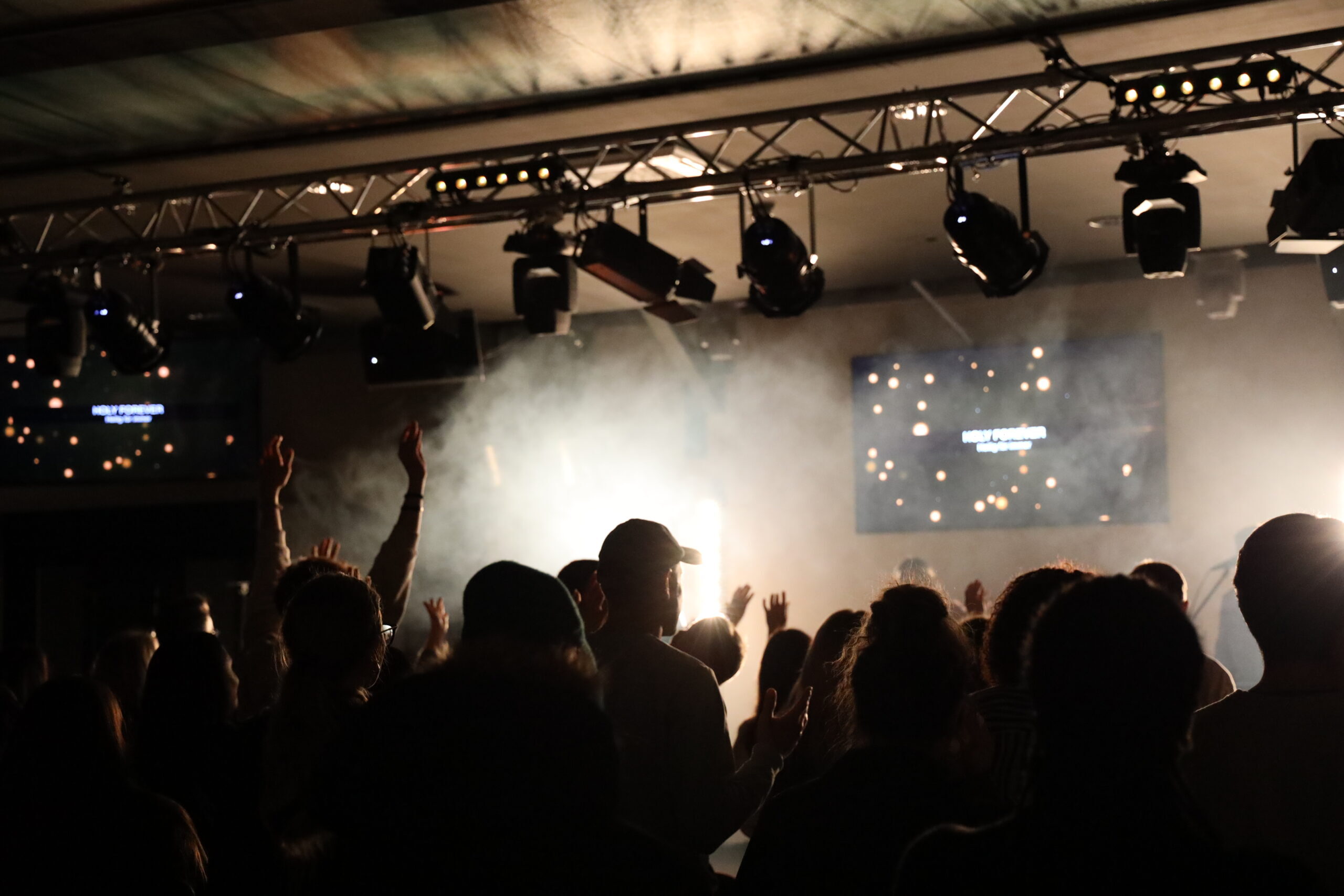 Menschen vor Bühne bei Konzert, einige mit erhobenen Armen, Lichteffekte im Hintergrund.
