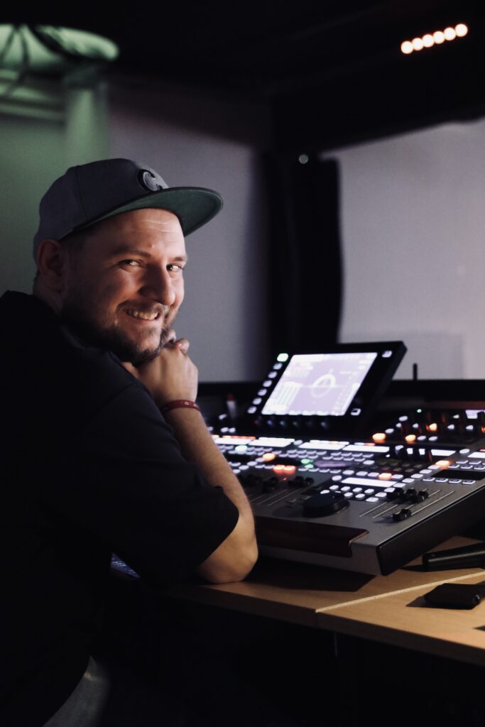 Mann mit Cap sitzt an Mischpult im Tonstudio, lächelt in die Kamera.