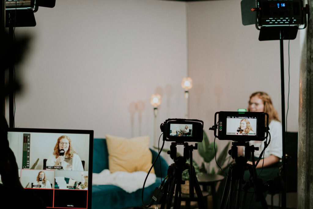 Ehefrau im Videochat während einer Live-Übertragung, auf einer Couch mit Kissen, in Hintergrund Pflanzen und Stehlampen, Kamera- und Lichttechnik im Einsatz, professionelle Aufnahme für Online-Gottesdienst oder Event.