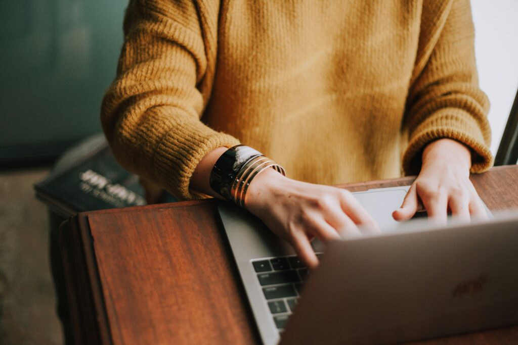 Alt Text: Person tippt auf einem Laptop, trägt einen goldfarbenen Pullover und eine große Armreifen, sitzt an einem Holzschreibtisch.