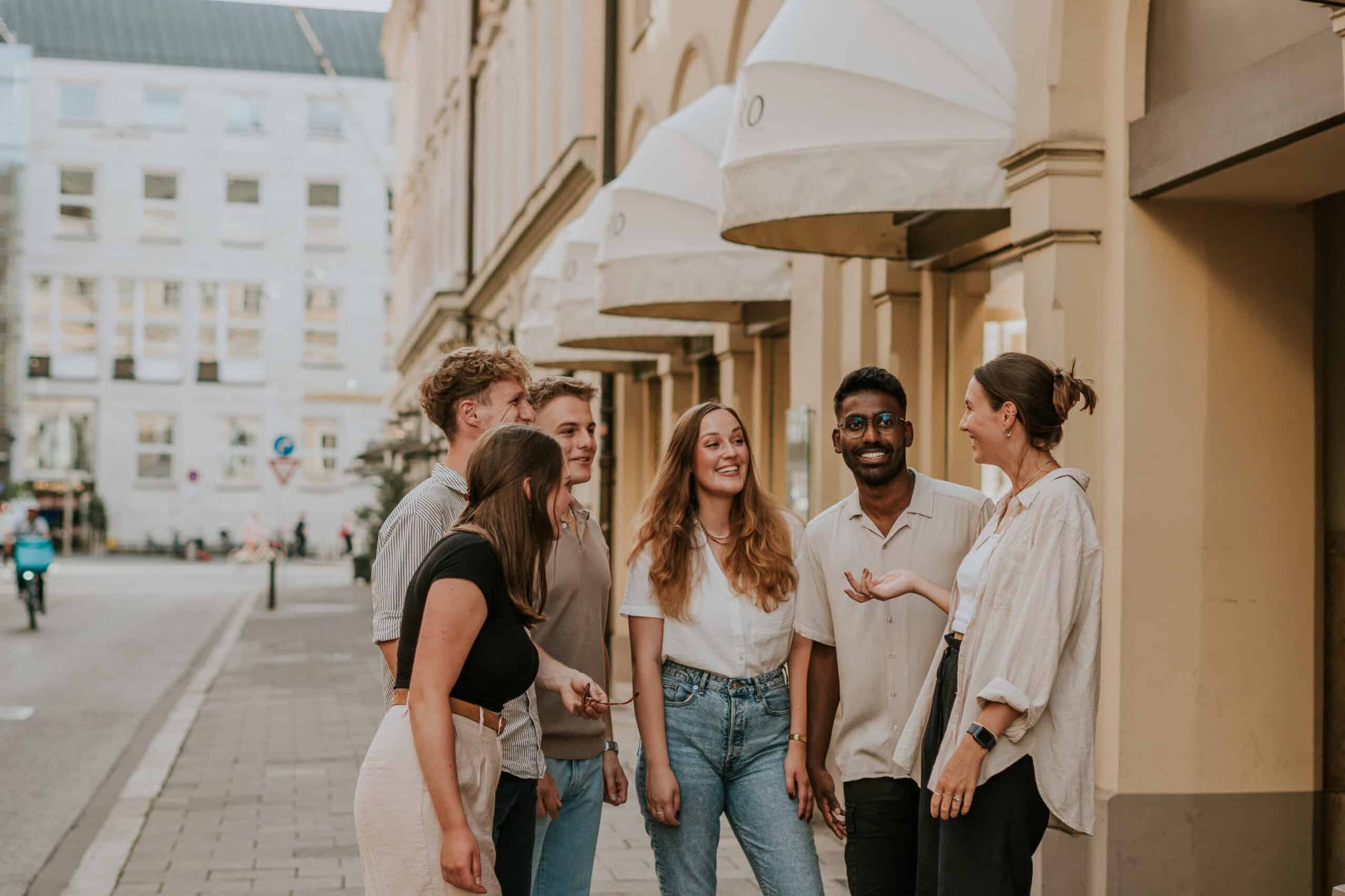 Menschen in einem Gespräch auf der Straße vor einem Gebäude mit Überdachungen, freundliche Atmosphäre, urbanes Umfeld, diverse Gruppe, moderne Kleidung, Stadtleben, soziale Interaktion, positive Stimmung, multikulturell.