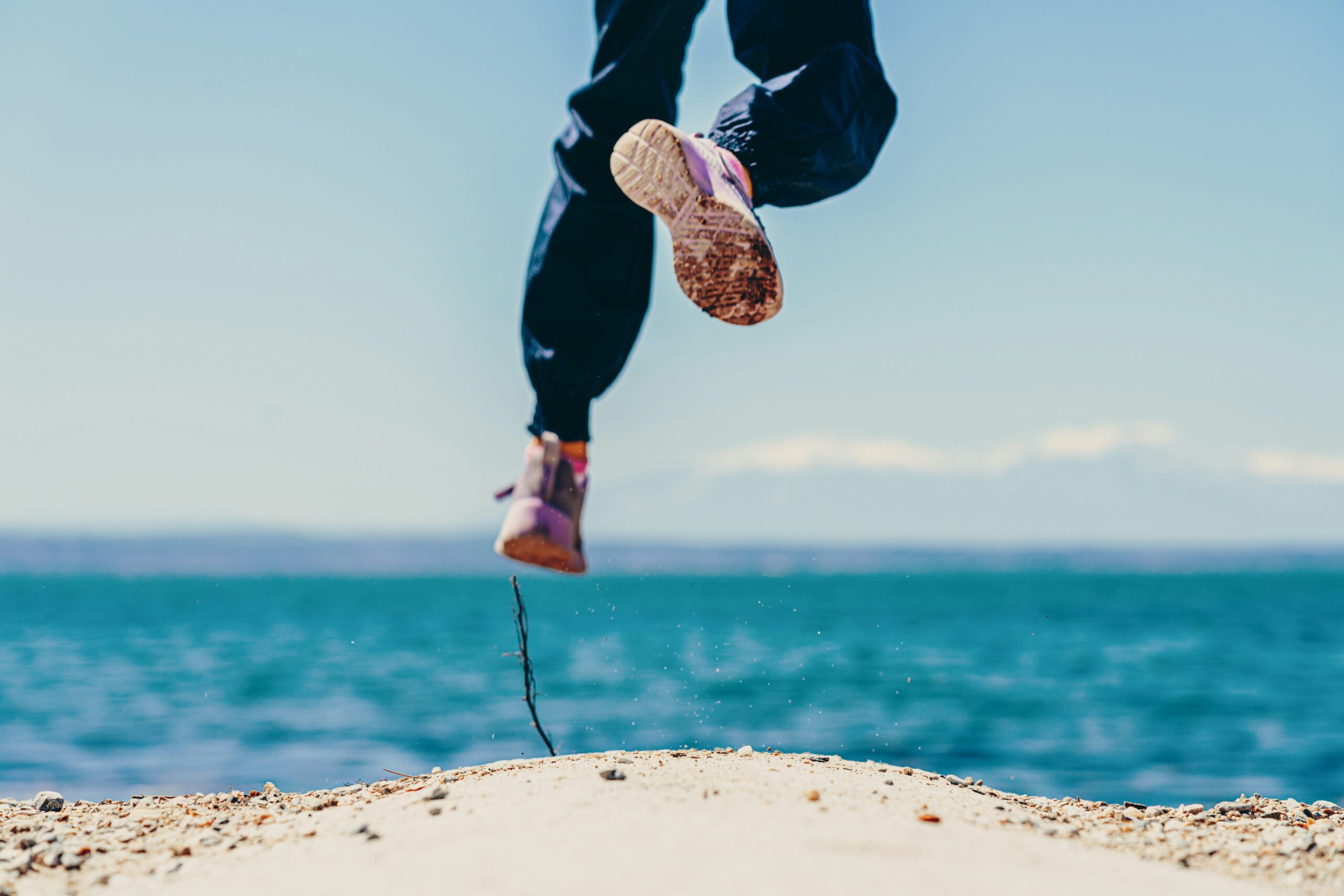 Person springt am Strand, Blick auf die Füße und das Meer im Hintergrund.