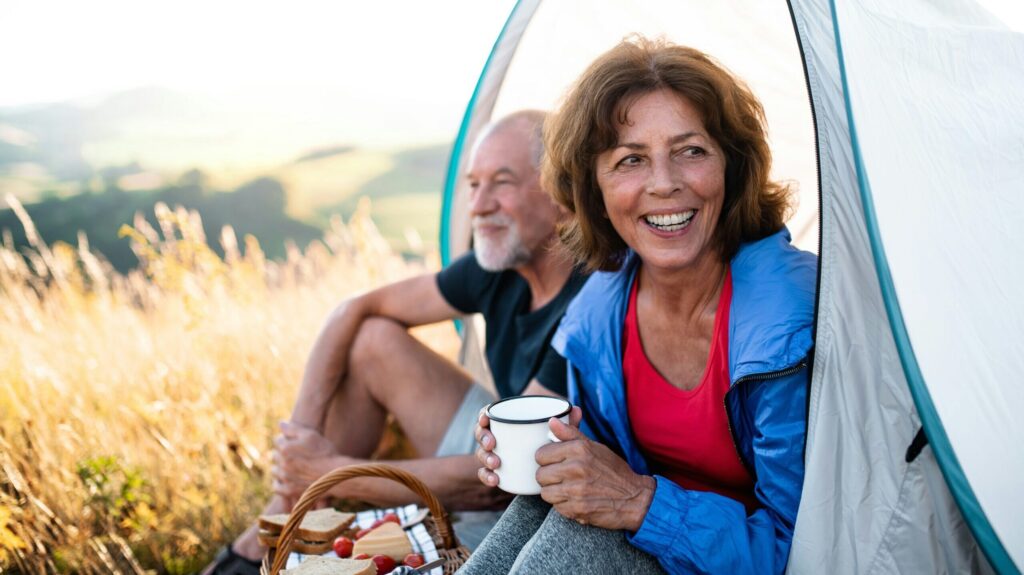 Frau lächelnd mit Becher vor Zelt, Mann im Hintergrund, Natur, Camping.