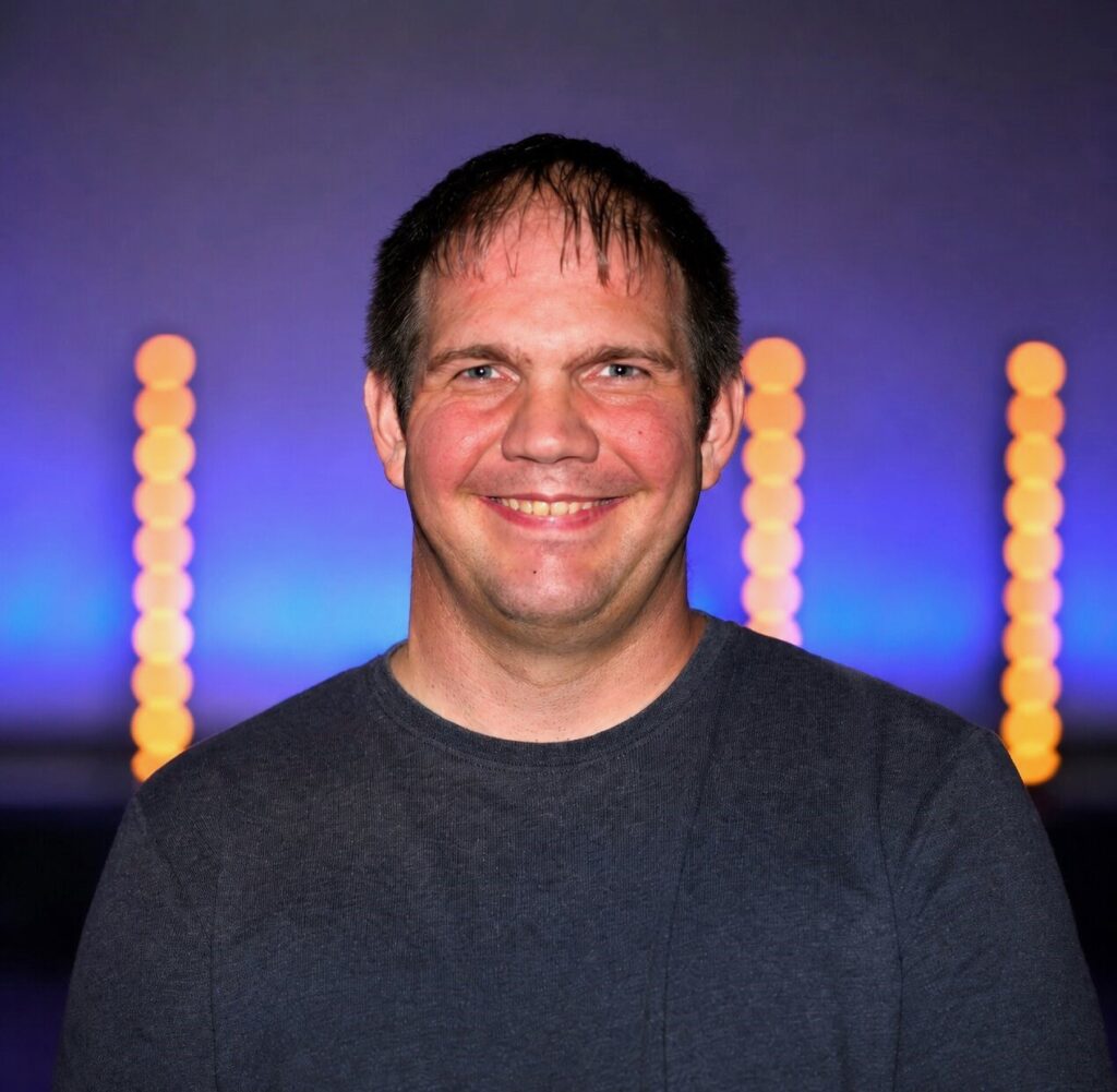 Mann mit dunklem T-Shirt vor bunten Lichtern im Hintergrund.