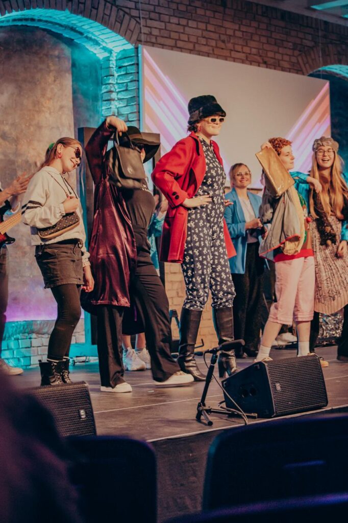 Mehrere Menschen in bunten Kostümen auf einer Bühne, einige tragen Hüte und Sonnenbrillen.