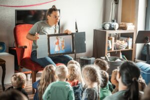 Lehrerin zeigt Kindern eine Illustration auf einem Bildschirm, die eine Figur mit einem roten Umhang.