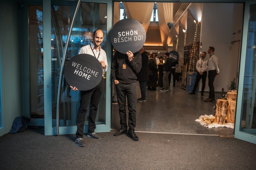 Zwei Männer stehen an der Eingangstür, einer hält ein Schild mit "Welcome Home", der andere mit "Sch.
