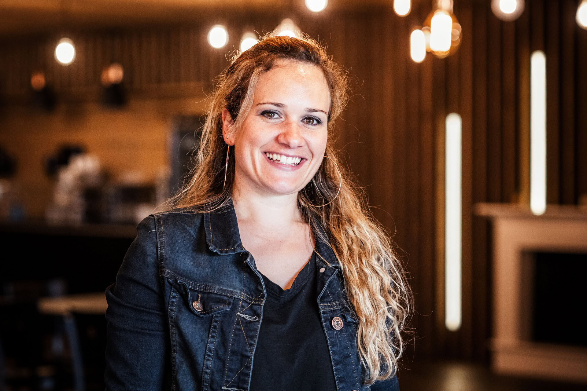 Frau mit lockigem Haar und dunkler Jacke lächelt in einem Restaurant mit warmem Licht.