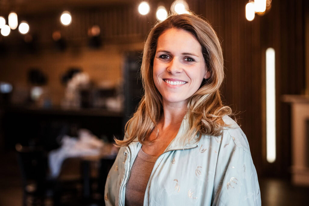 Eine Frau mit braunen, gewellten Haaren lächelt in einem Café.