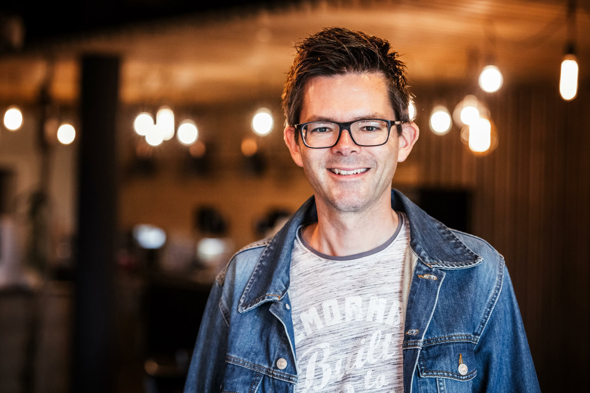 Mann mit Brille, kurzhaarig, trägt Jeansjacke und graues T-Shirt, lächelt.
