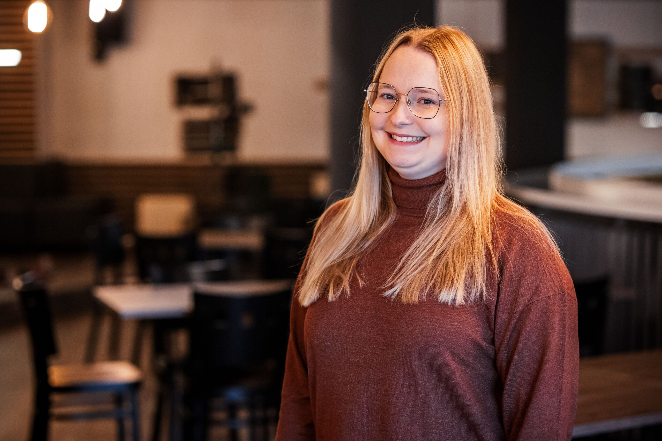 Eine junge Frau mit langen, roten Haaren und Brille lächelt in einem Café.