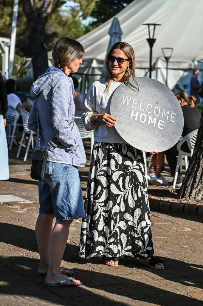Zwei Frauen im Gespräch, eine hält ein "Welcome Home" Schild, im Hintergrund Zelte und Bäume.