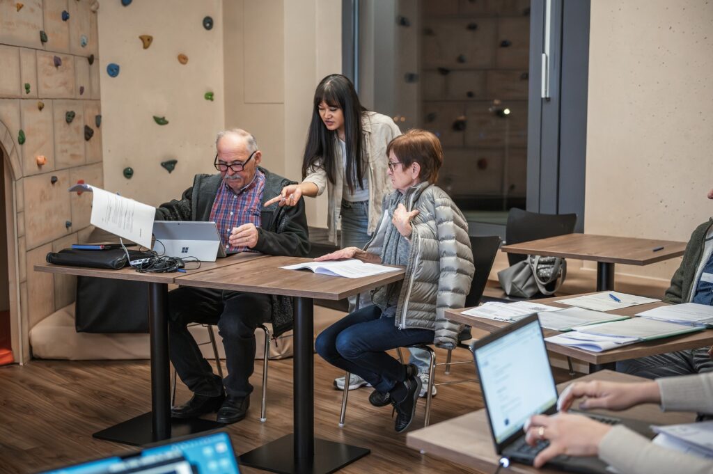 Drei Personen arbeiten an einem Tisch, zwei sitzen, eine steht, mit Laptops und Dokumenten.