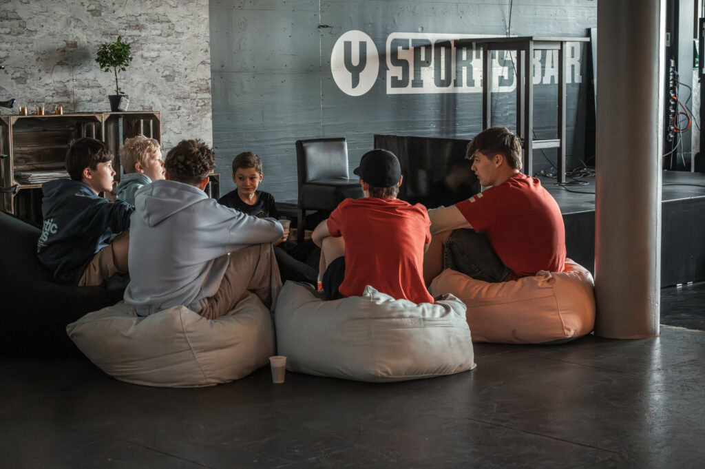 Sechs junge Menschen sitzen auf Sitzsäcken im Fitnessstudio, unterhalten sich.