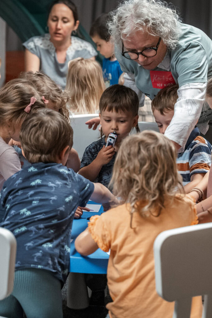 Erwachsene und Kinder bei einer Aktivität, Kinder sitzen um einen Tisch, eine Frau zeigt auf einen G.