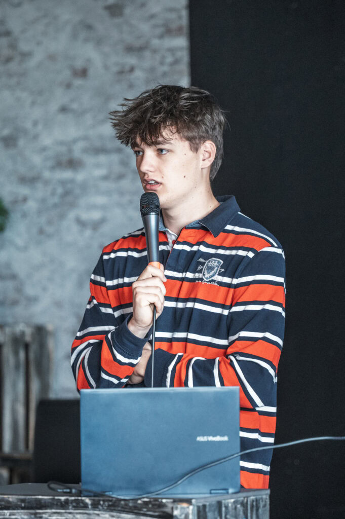 Ein junger Mann hält ein Mikrofon in der Hand, steht vor einem Laptop.