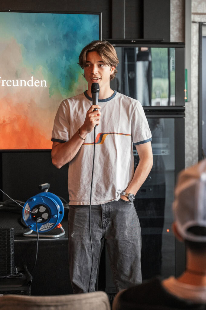 Junger Mann hält Mikrofon und spricht vor Publikum in einem Raum.