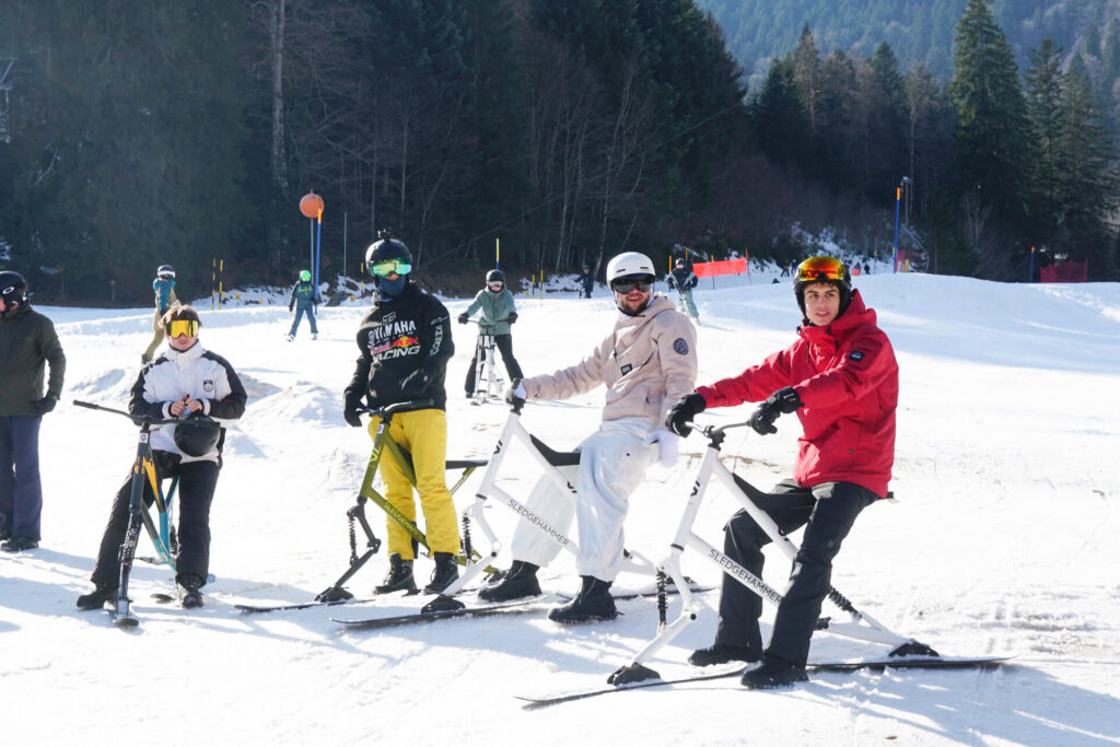 Fünf Personen mit Schneeschuhen auf verschneitem Berg, im Hintergrund Bäume und Skilifte.