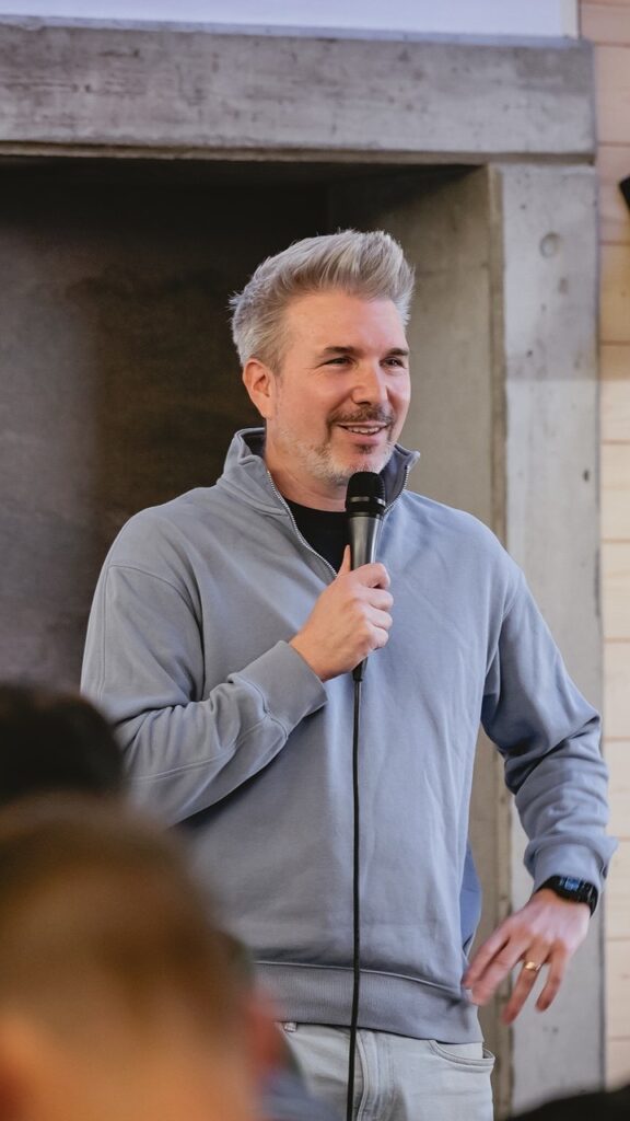 Mann mit grauem Haar und grauem Pullover spricht in Mikrofon.