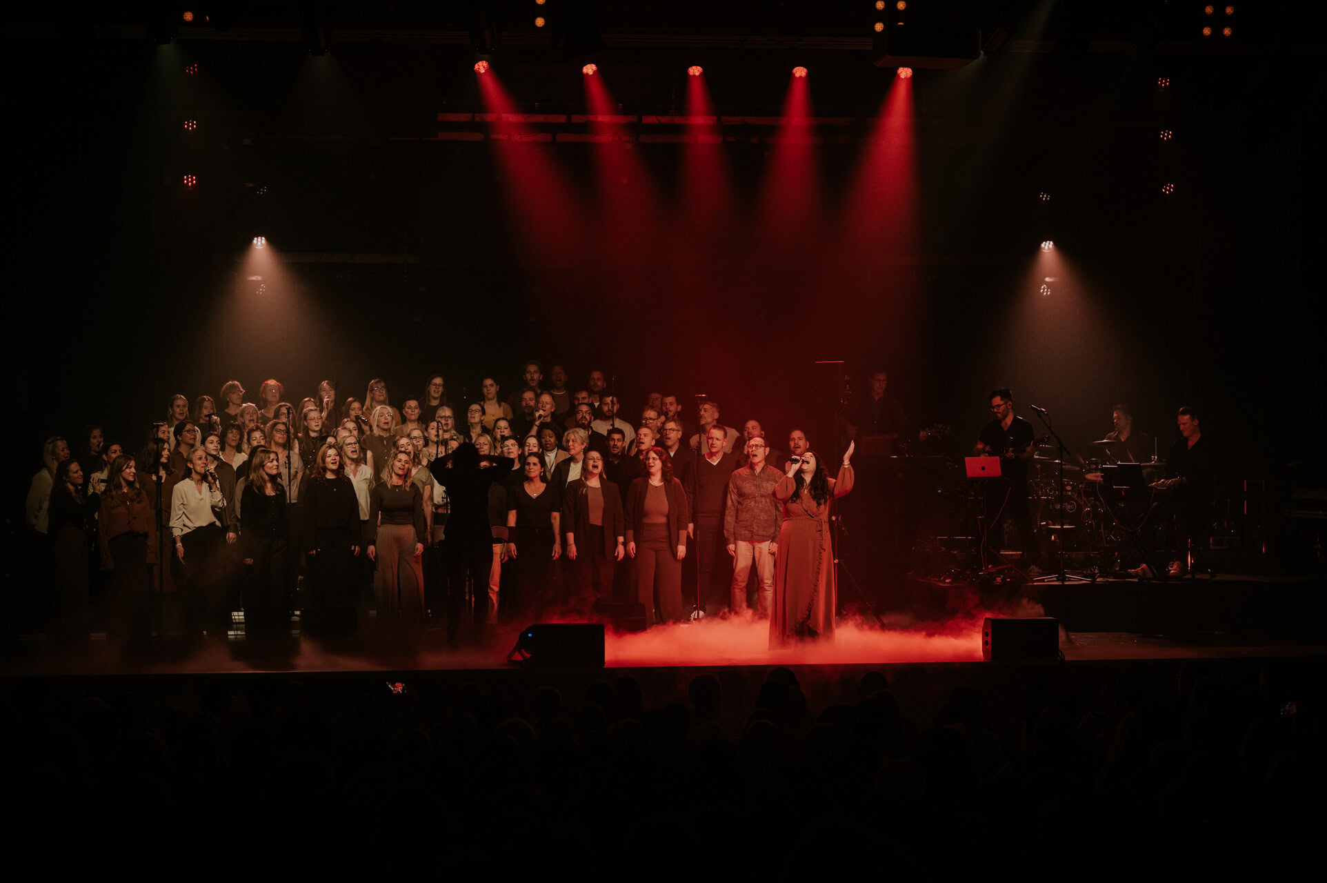 Große Gesangsgruppe auf einer Bühne bei einem Konzert, mit Musikern und Bühnenbeleuchtung.