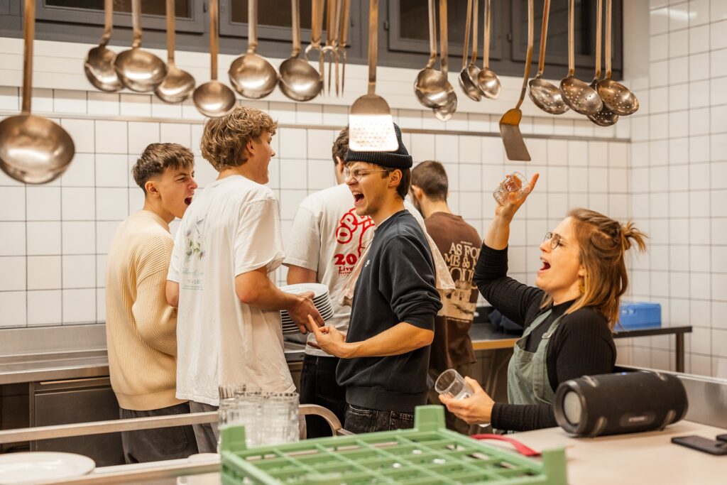Menschen in einer Küche unter einer Reihe von Kochlöffeln und Pfannen.