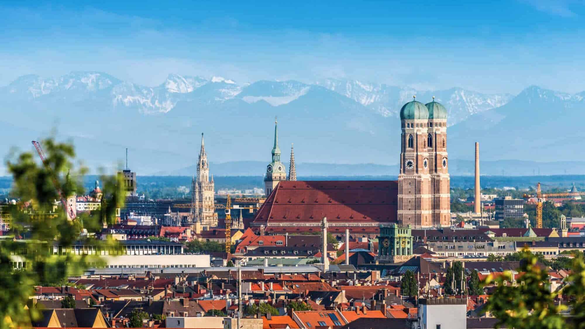 Hochwertige Panoramaaufnahme von München mit dem bekannten Rathaus und den Alpen im Hintergrund, betont die lebendige Stadt mit bayerischer Kultur und religiöser Tradition.