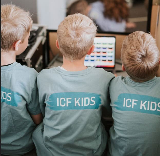 Alt-Text: Drei Kinder in ICF-Kids-Shirts sitzen beim Lernen an Computern im Gemeinschaftsraum des ICF München, was eine freundliche und kreative Lernumgebung zeigt.