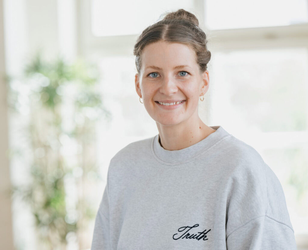 Helle junge Frau mit grauem Sweatshirt, lächelnd vor Fenster im Hintergrund.