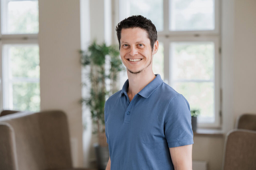 Mann in blauem Polo-Shirt steht in einem hellen Wohnzimmer mit großen Fenstern.
