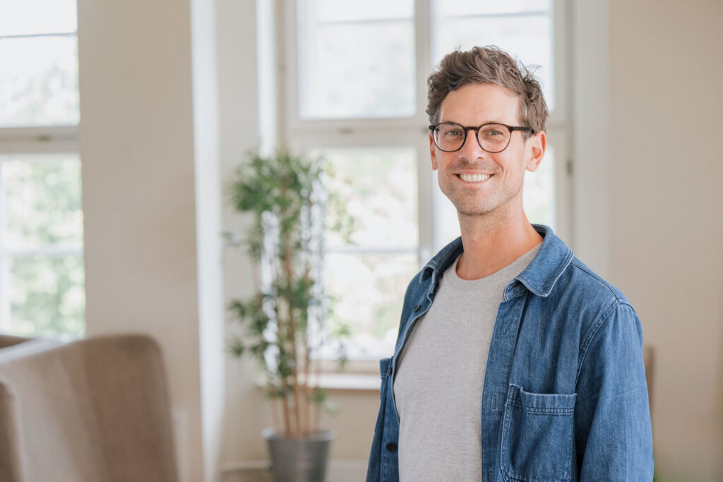 Mann mit Brille, grauem T-Shirt und Jeans in einem hellen Raum.