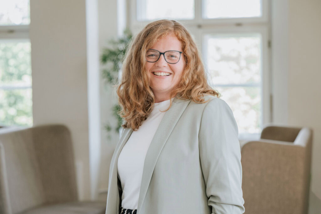 Frau mit Brille und lockigem, rotem Haar lächelt in einem hellen Raum.