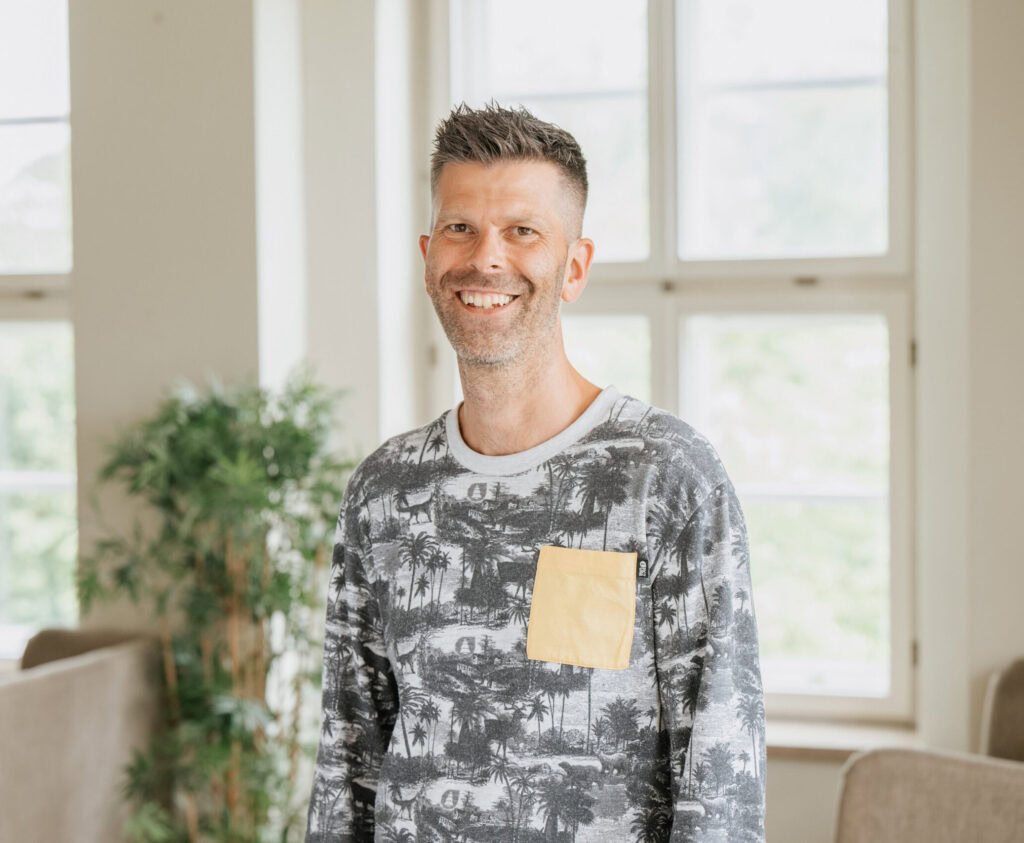 Ein Mann steht in einem Raum mit großen Fenstern im Hintergrund.