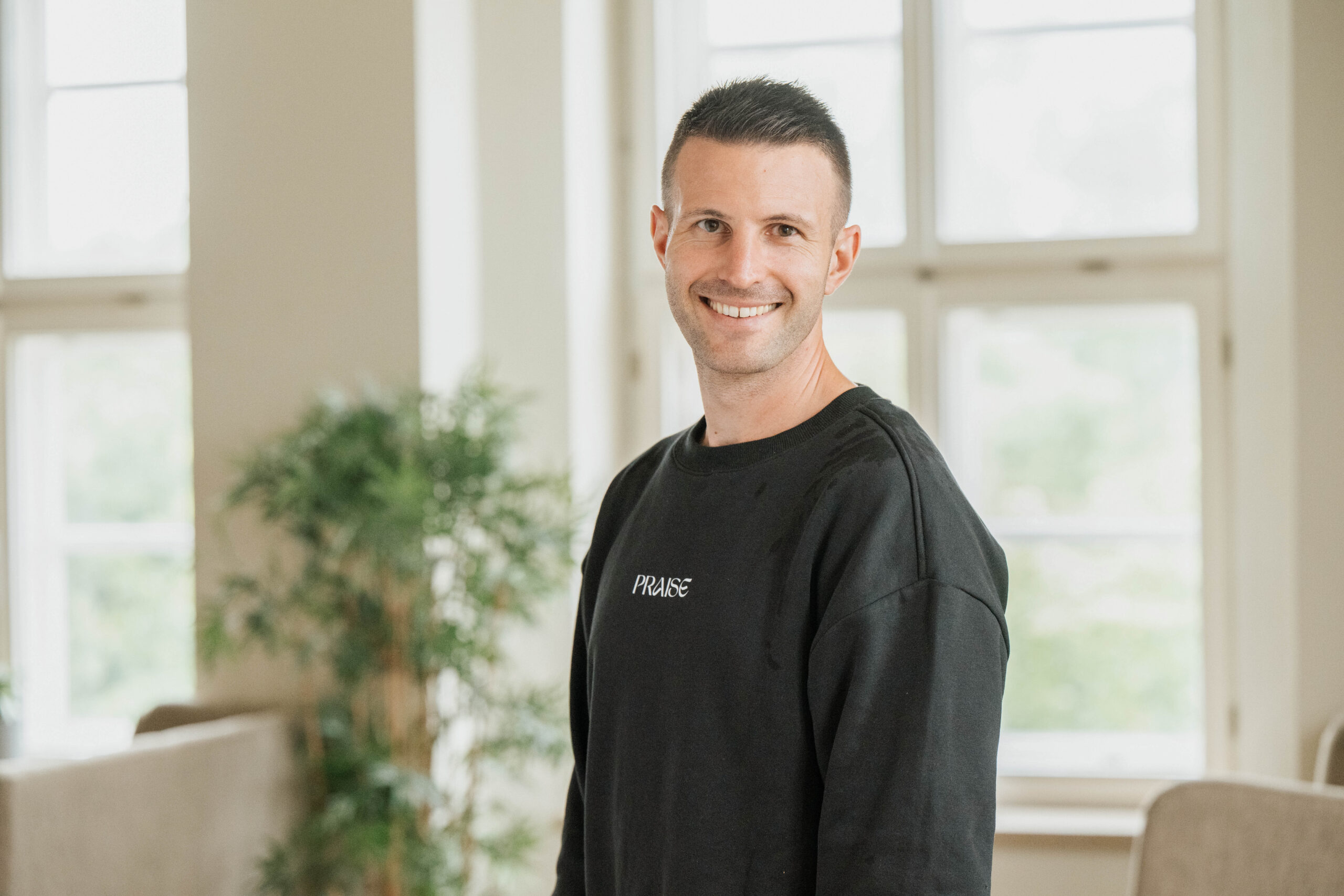 Ein Mann in schwarzem Pullover steht in einem hellen Raum mit großen Fenstern.