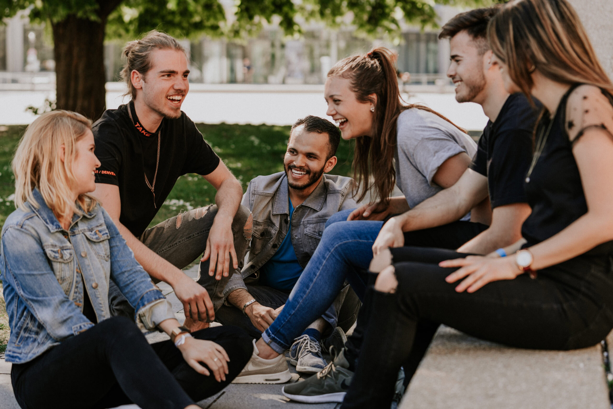Mehrere junge Menschen sitzen im Kreis auf dem Boden im Park und sprechen miteinander.