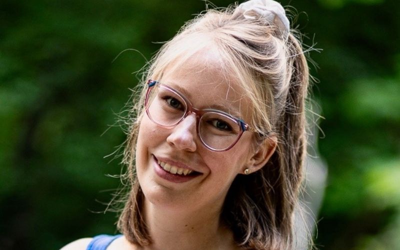 Junge Frau mit Brille lächelt im Park, grüne Bäume im Hintergrund.