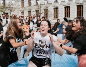 Mehrere Menschen lachen und feiern im Freien, eine Person wird in einem Wasserbecken nass.