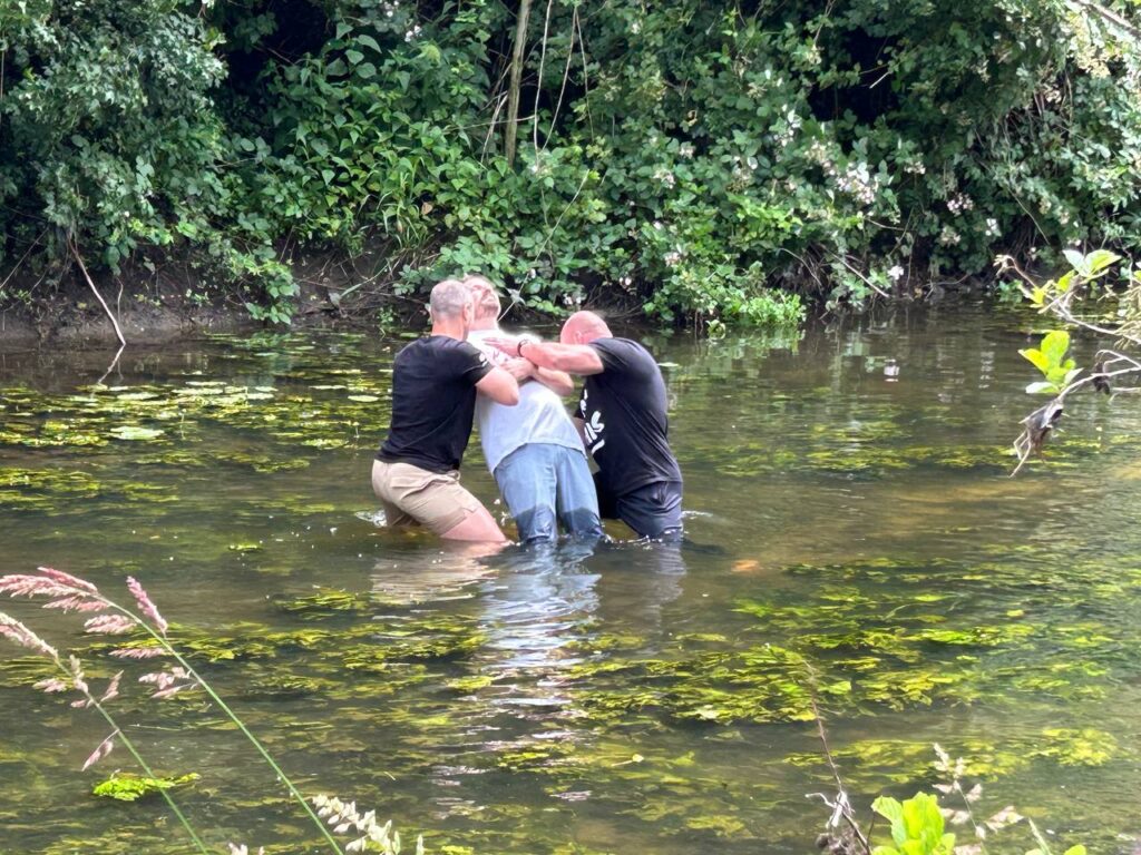 Drei Männer stehen im Fluss, halten sich an den Armen und unterstützen eine Person beim Wasser.