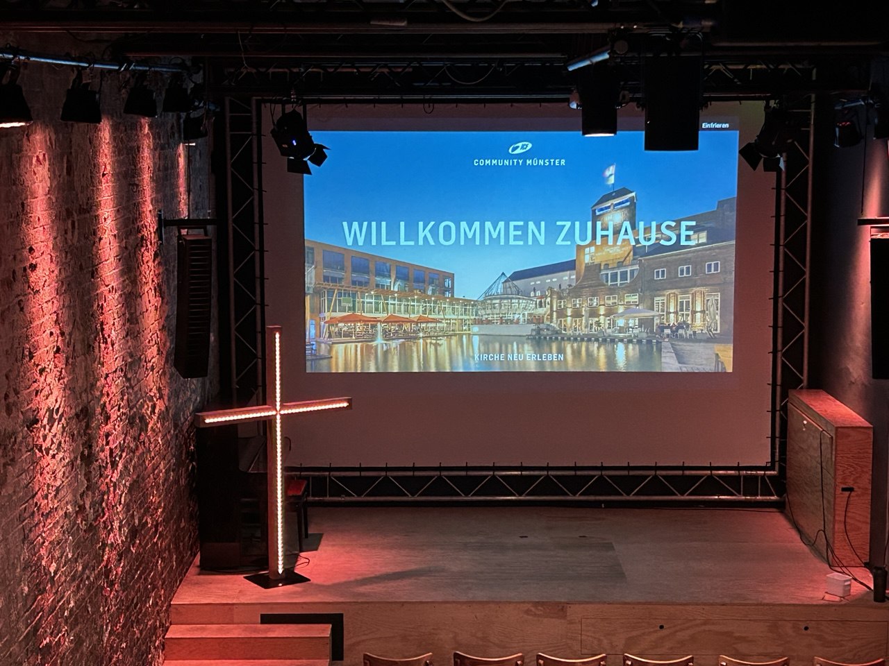 Leeres Bühnenbild mit Projektorbildschirm, der "Willkommen Zuhause" zeigt.