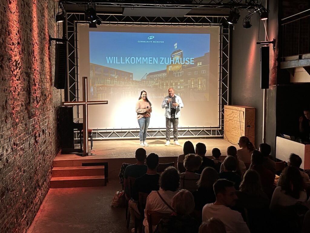 Zwei Personen stehen auf einer Bühne vor einem Publikum, im Hintergrund ein Bildschirm mit "WILLKOMM.