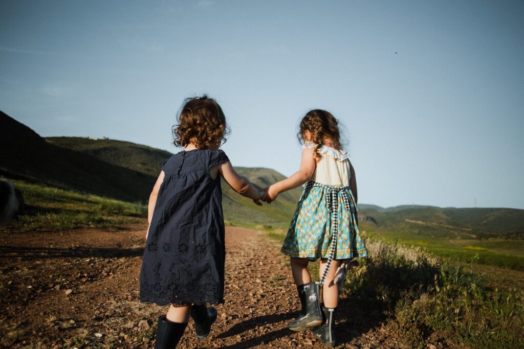 Alt-Text: Zwei Kinder halten Händchen beim Spaziergang auf einem Feldweg in den Bergen, umgeben von grüner Landschaft, klarer Himmel und Natur, Familienaktivität, kindgerechte Erkundung.