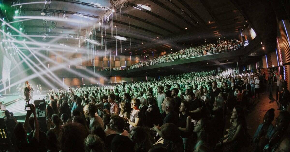 A large audience seated in a concert hall with stage lights and a performer on stage.