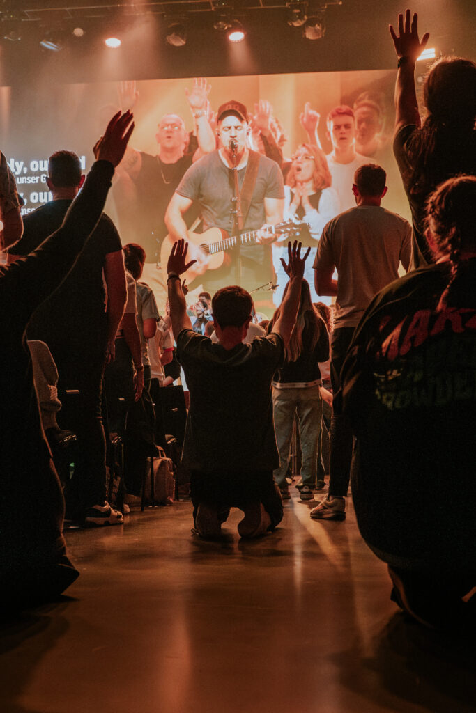 Group of people raising hands during a worship service with a large screen showing a musician playin.
