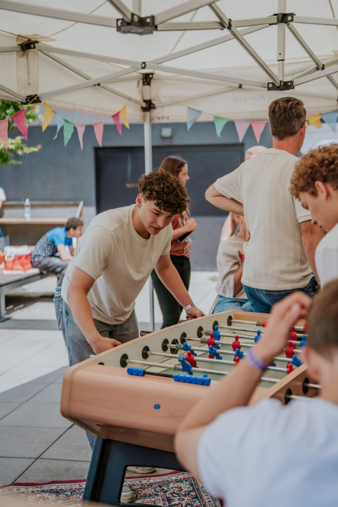 A group of young people playing foosball under a white canopy tent decorated with colorful pennant b.